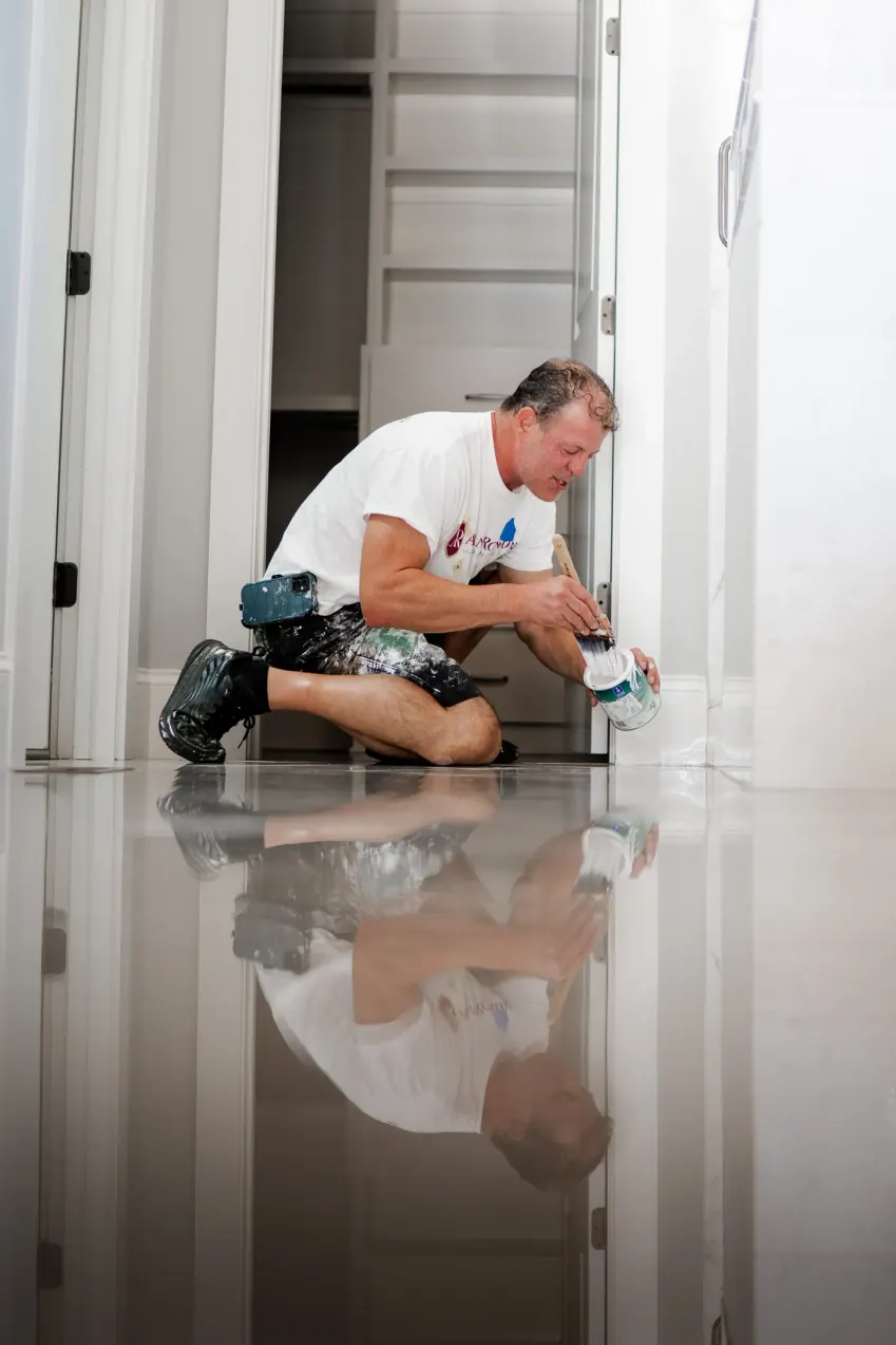 Alan Robinson kneeling on a bright bathroom floor, painting the baseboard by hand, interior painting by hand for Fairview homes