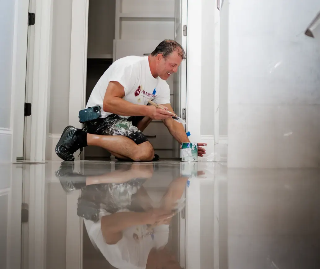 Alan Robinson kneeling in a doorway, brushing paint onto a door casing beside an open paint can
