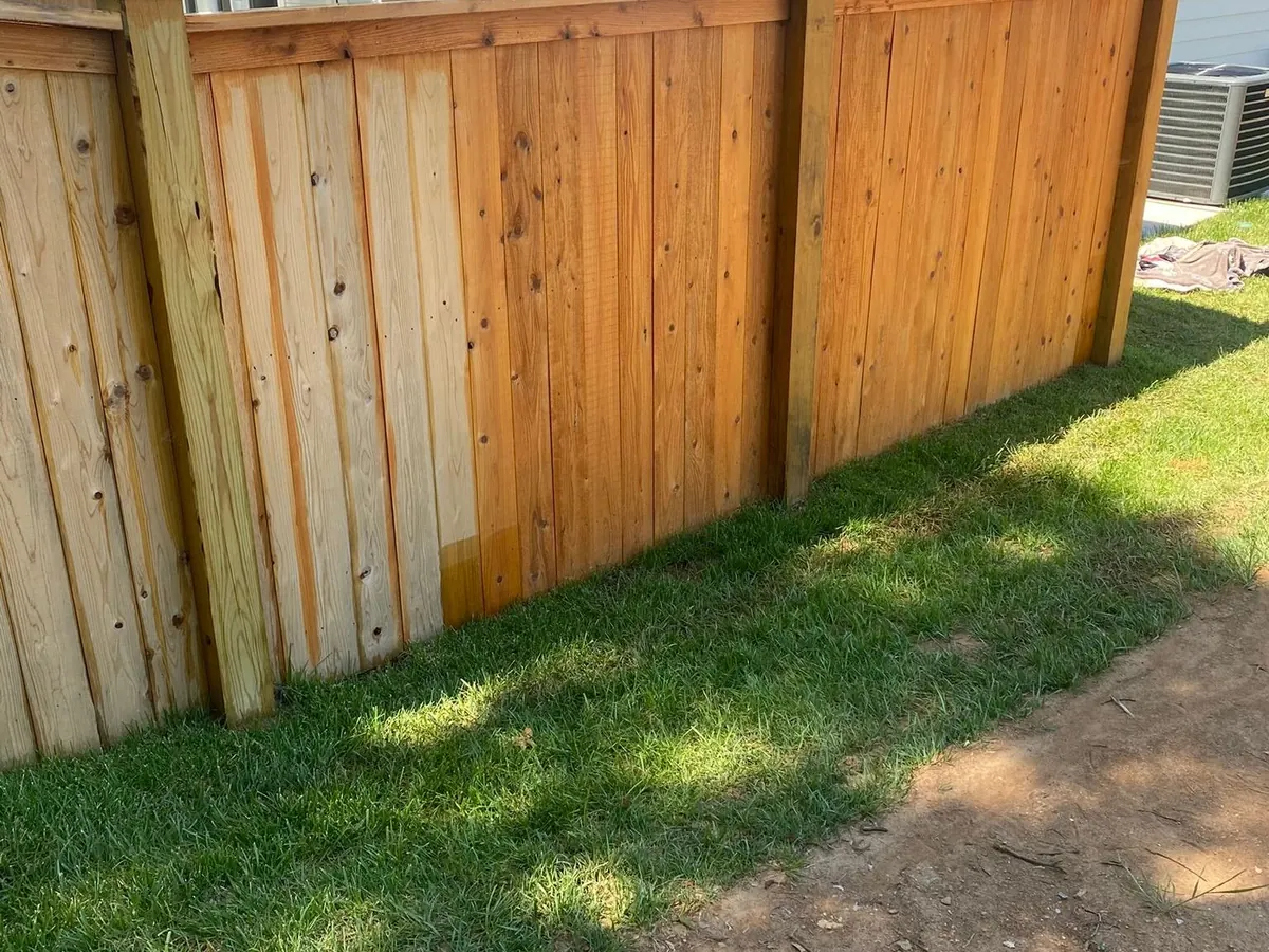 Privacy fence mid-stain, fresh cedar tone beside raw boards along a side yard, part of the fence staining we do for Bellevue homes