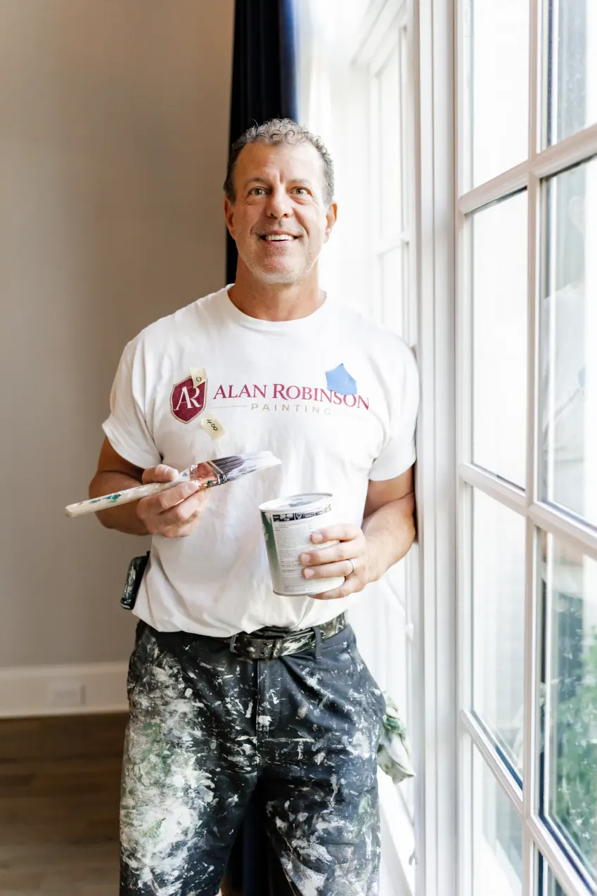 Alan Robinson smiling beside a tall window with a paintbrush and a cup of paint, interior painting by hand for Nashville homes